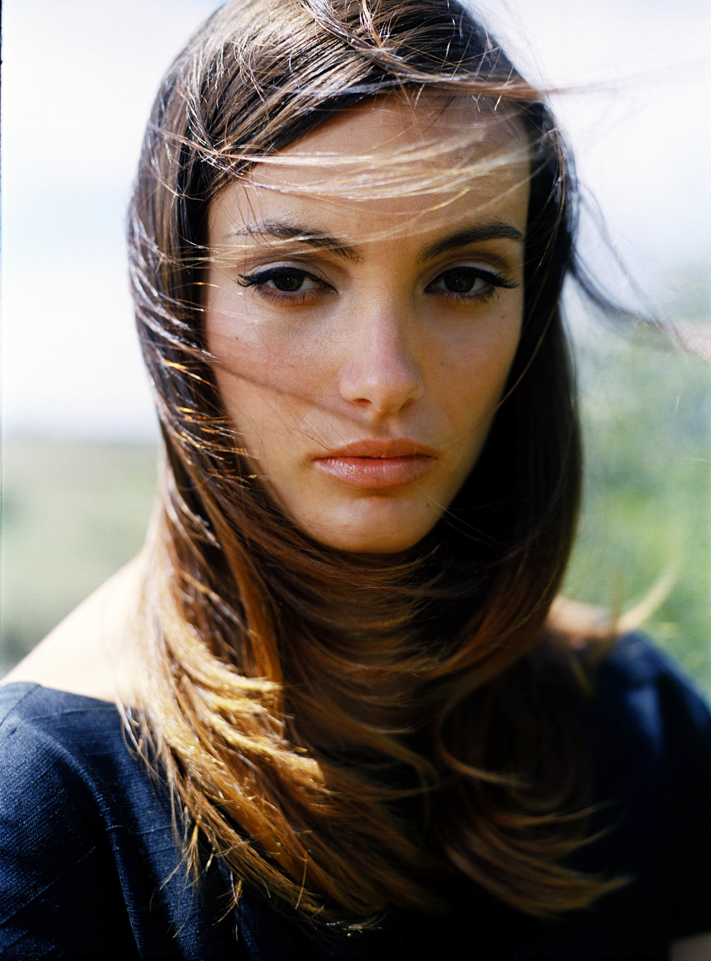 Portrait of a young woman with long brown hair blowing in the wind, wearing a dark top, against a soft blurred natural background.