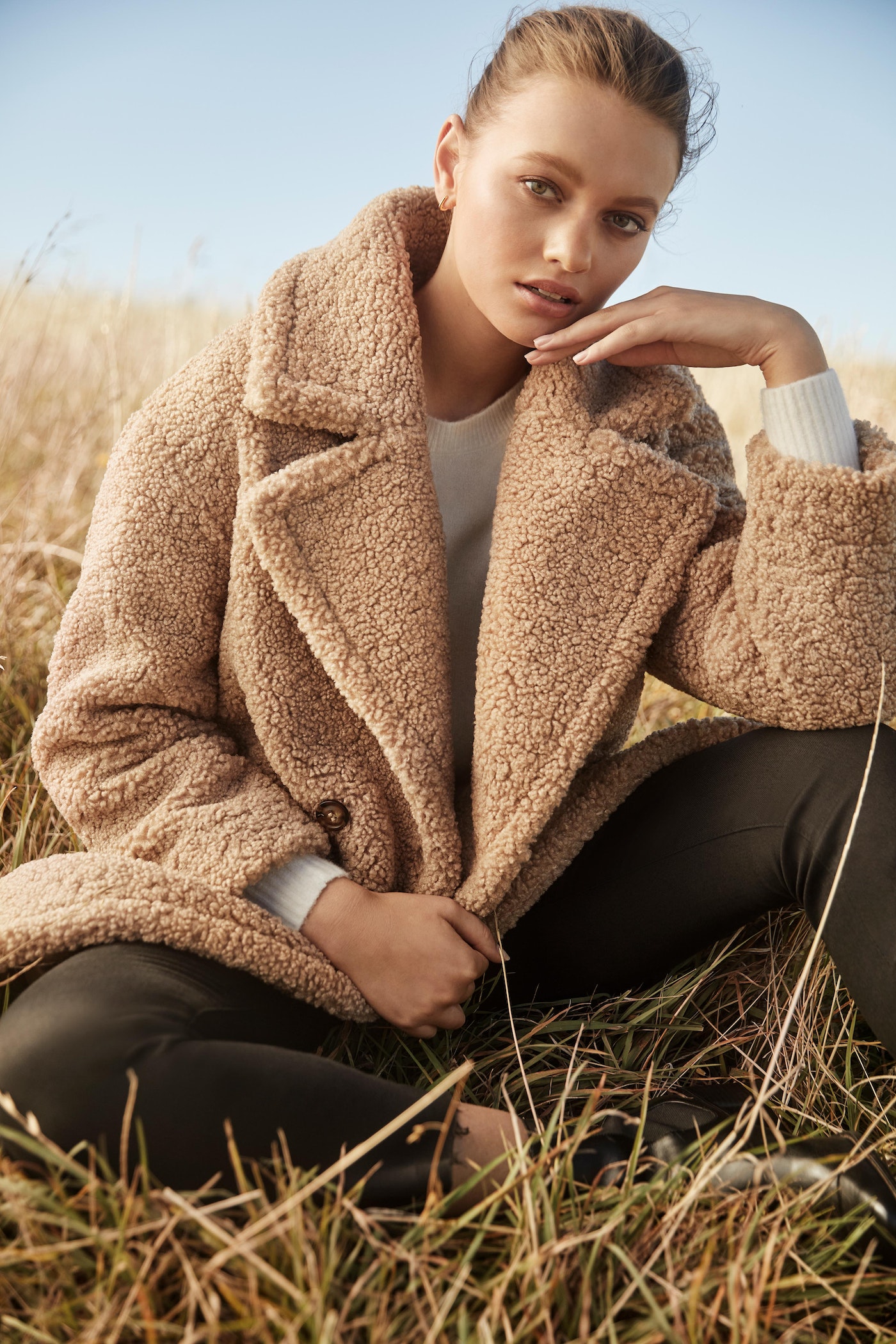 Woman poses in a teddy coat in a field of golden grasses under a bright sky. Soft lighting and natural setting highlight the fashion.