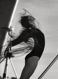 Monochrome portrait of a dancer in a black bodysuit with sheer flowing veil fabric captured in motion against an outdoor sky
