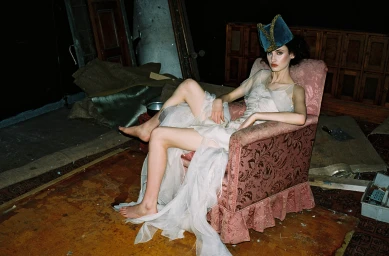 Woman in sheer white dress and blue velvet hat seated in an armchair in an untidy room