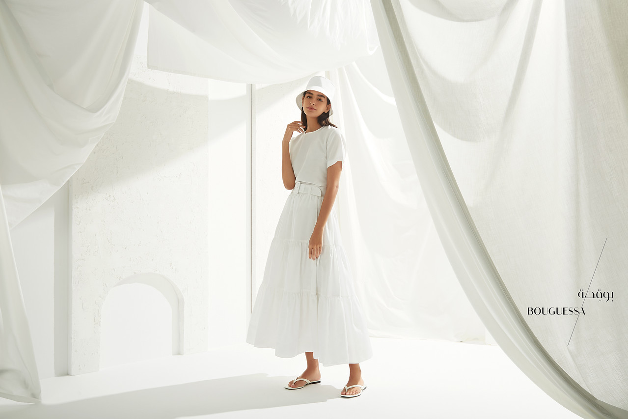 Fashion shot of a woman wearing a white dress and hat, posing indoors with white draped fabrics and minimalist decor, bright, airy aesthetic.