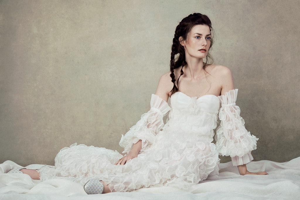 Woman in white strapless dress with braided hair sits indoors against neutral wall, captured in a studio portrait, conveying elegance and artistry.