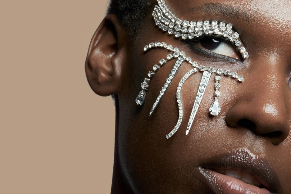 Close-up beauty shot of a Black woman's face with dramatic rhinestone embellishments around the eye in a studio setting.