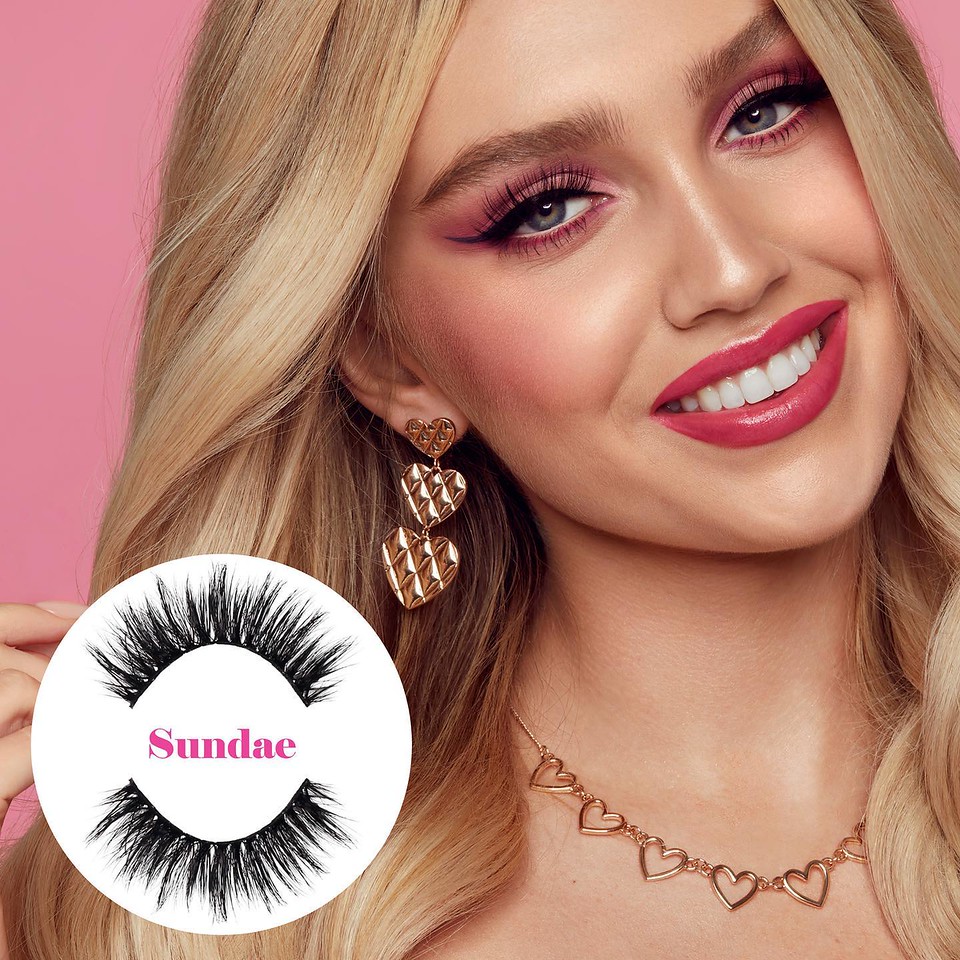Close-up shot of a smiling adult female with blonde hair, wearing heart-shaped earrings and necklace, and pink eye shadow, on a pink background.