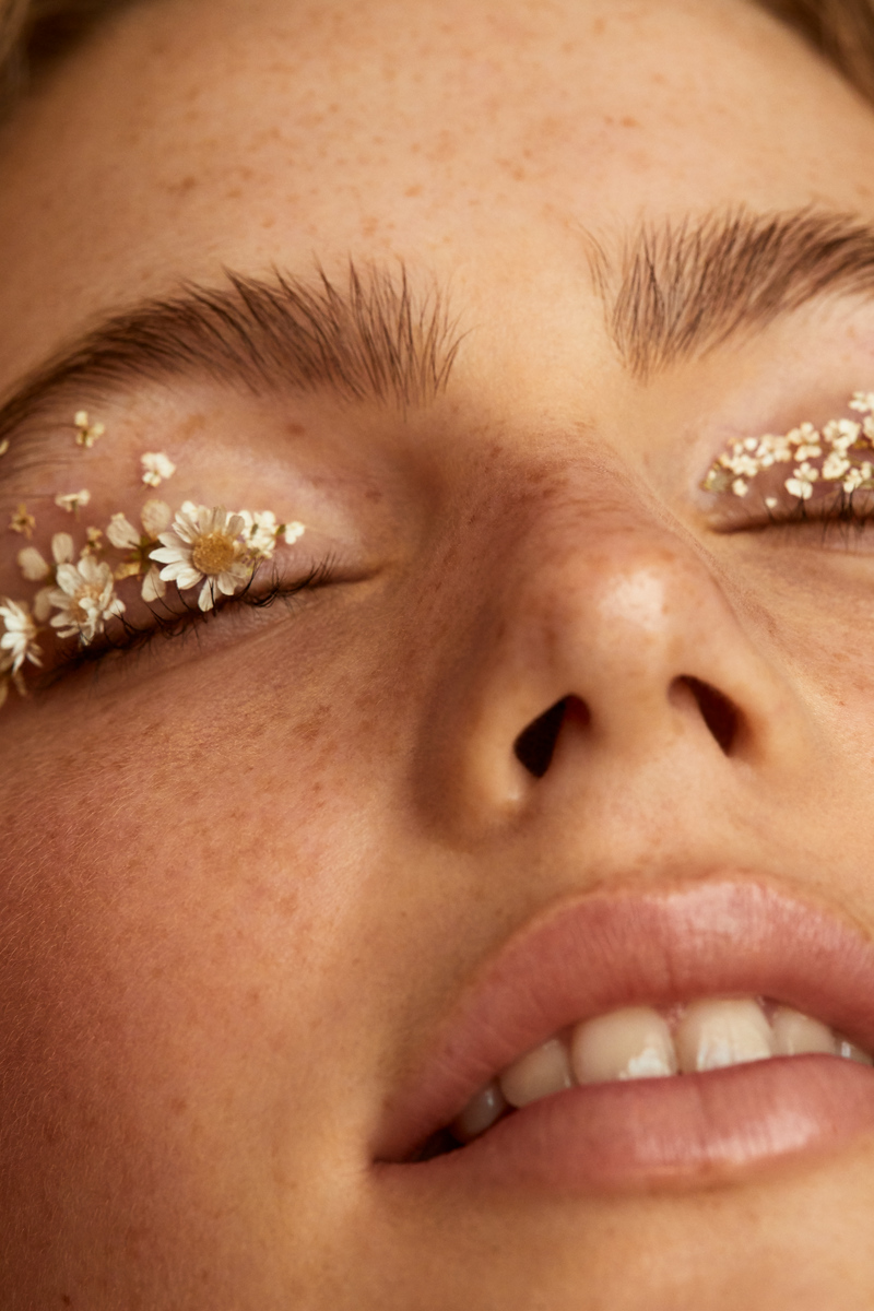 Close-up of a woman's face with freckles, her eyes closed and adorned with small white flowers on her eyelids, against a neutral background.