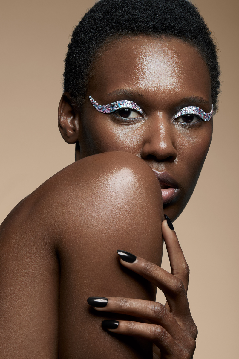 Close-up of a Black woman with glittery eye makeup, her hand near her shoulder, against a warm neutral backdrop. Focus on makeup and skin.