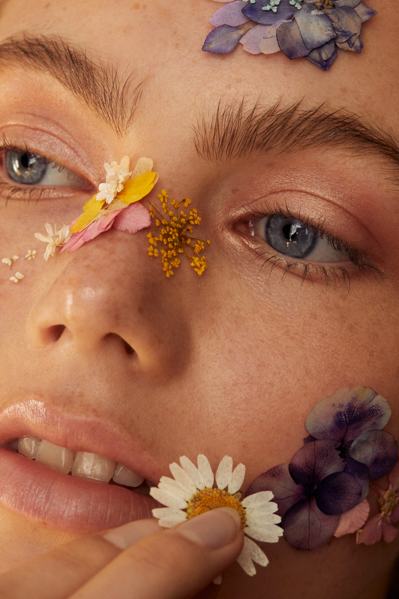 Close-up beauty shot of a young woman's face adorned with colorful flower petals and blossoms around her eyes, cheeks, and lips.