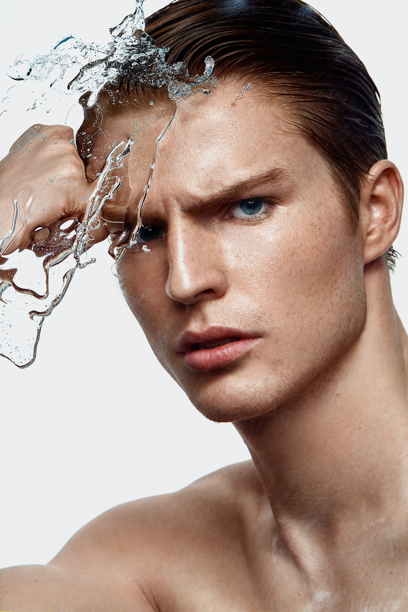 Dramatic portrait of a shirtless adult male with slicked-back hair, water splashing on his face against a neutral light background.