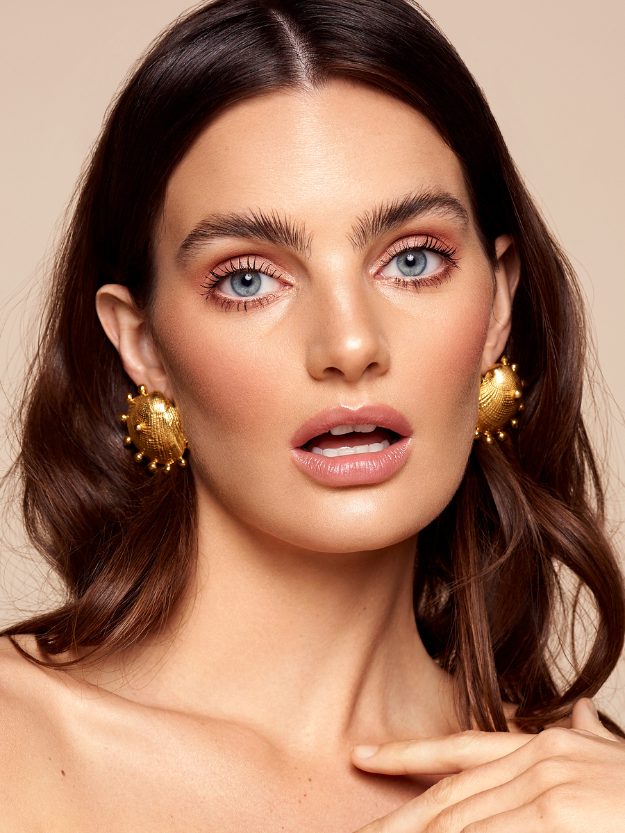 Close-up portrait of an adult woman with blue eyes, wearing large gold earrings, and light makeup against a neutral backdrop.