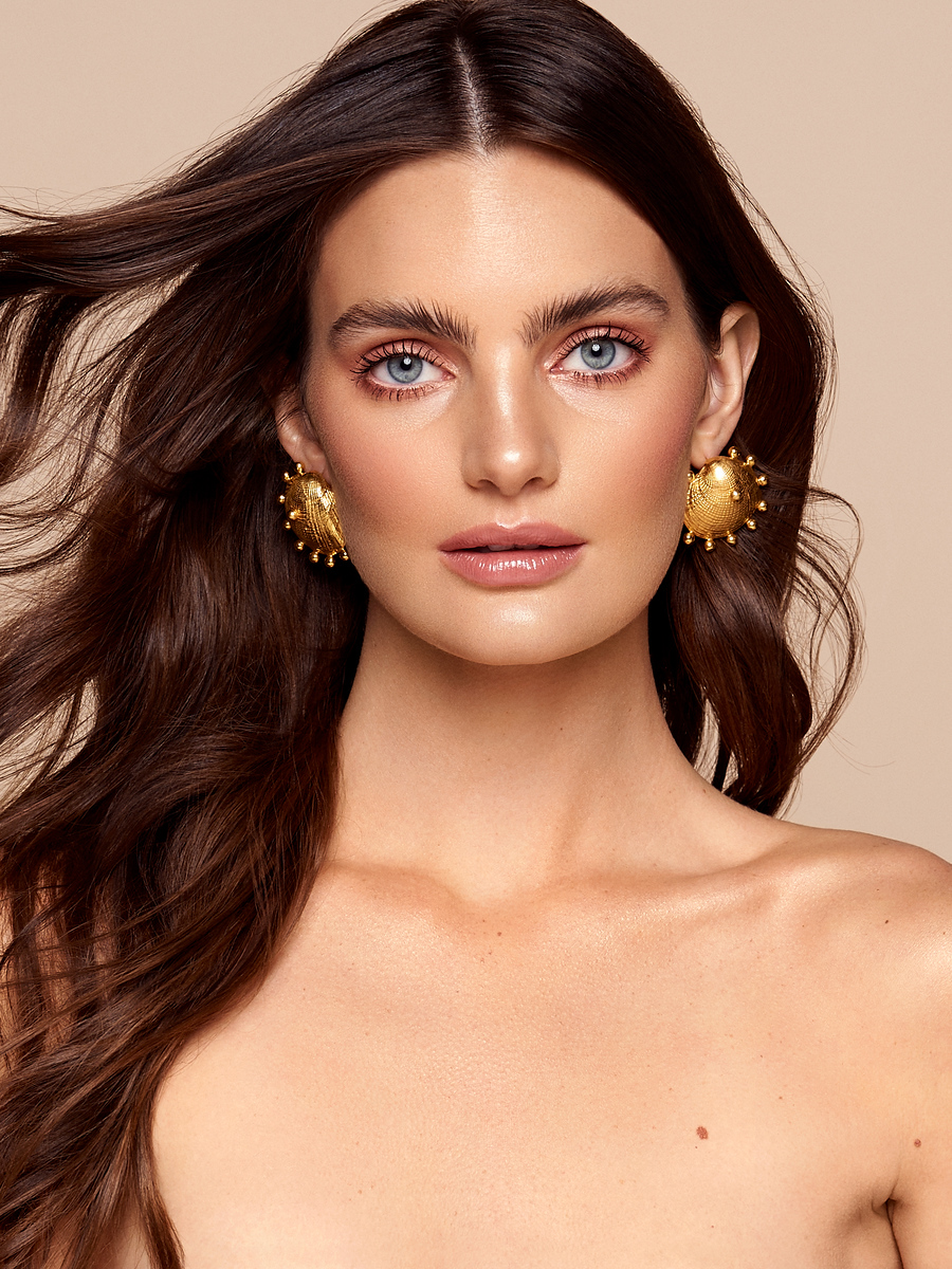 Close-up portrait of a woman with long brown hair, light eyes, wearing large gold earrings against a neutral beige backdrop.