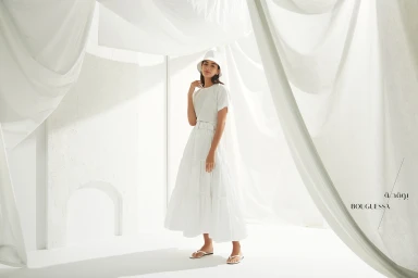 Model in white bucket hat, t-shirt, and tiered skirt standing against draped white fabric backdrop
