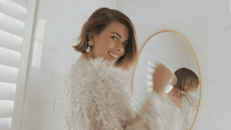 Smiling woman with short brown hair wearing a glittering fringed jacket in a bright, white bathroom with a round mirror.