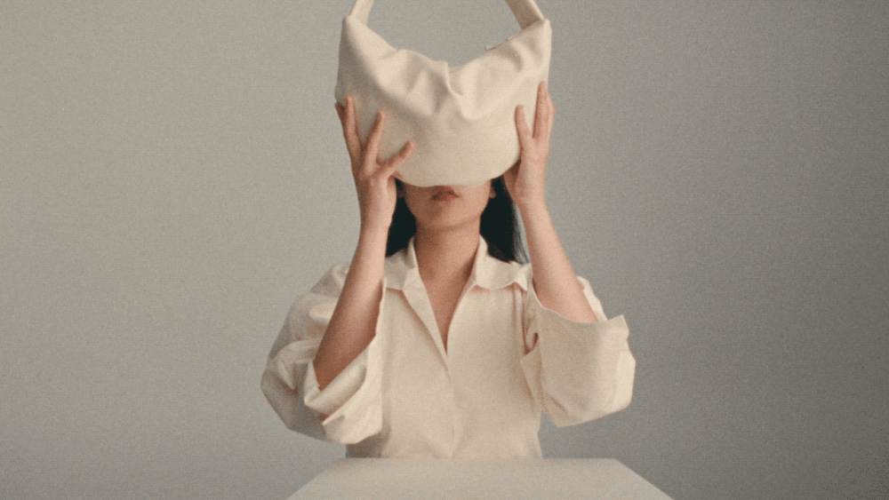 Portrait of Asian woman with a white handbag over her head in front of a gray background, posing with a slight smile. Fashion shot.