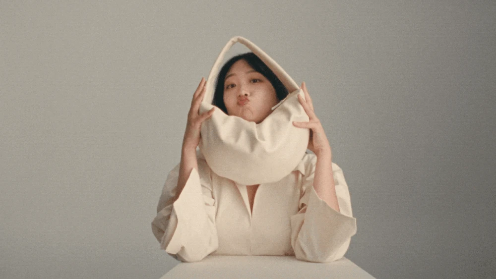 Portrait of Asian woman with a white handbag over her head in front of a gray background, posing with a slight smile. Fashion shot.
