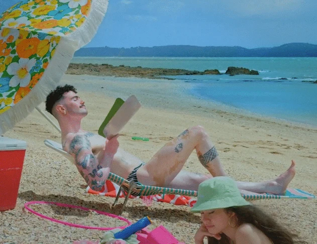 Adult male relaxes in a beach chair under a flowered umbrella while an adult female in a bucket hat sits nearby on a sandy beach.