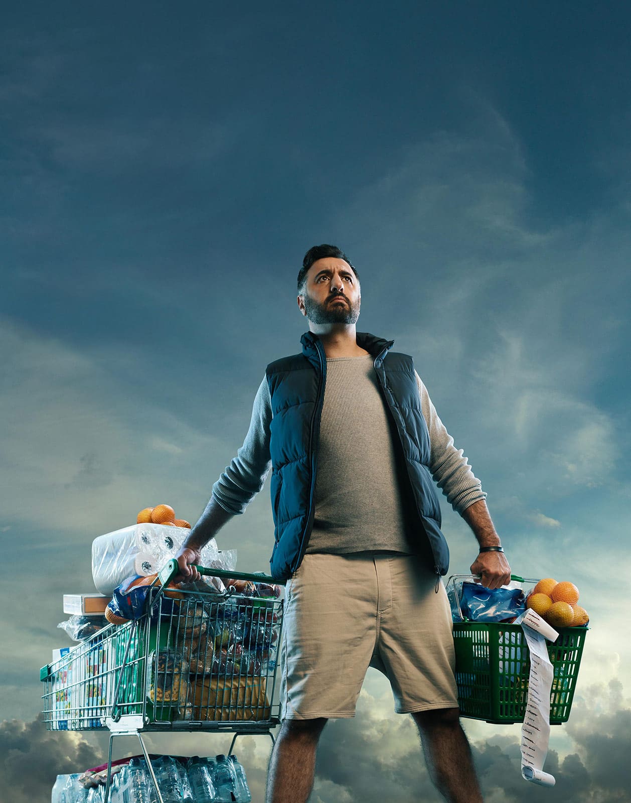 Bearded man poses heroically with a full shopping cart and basket against a dramatic sky backdrop, illuminated by studio lighting.