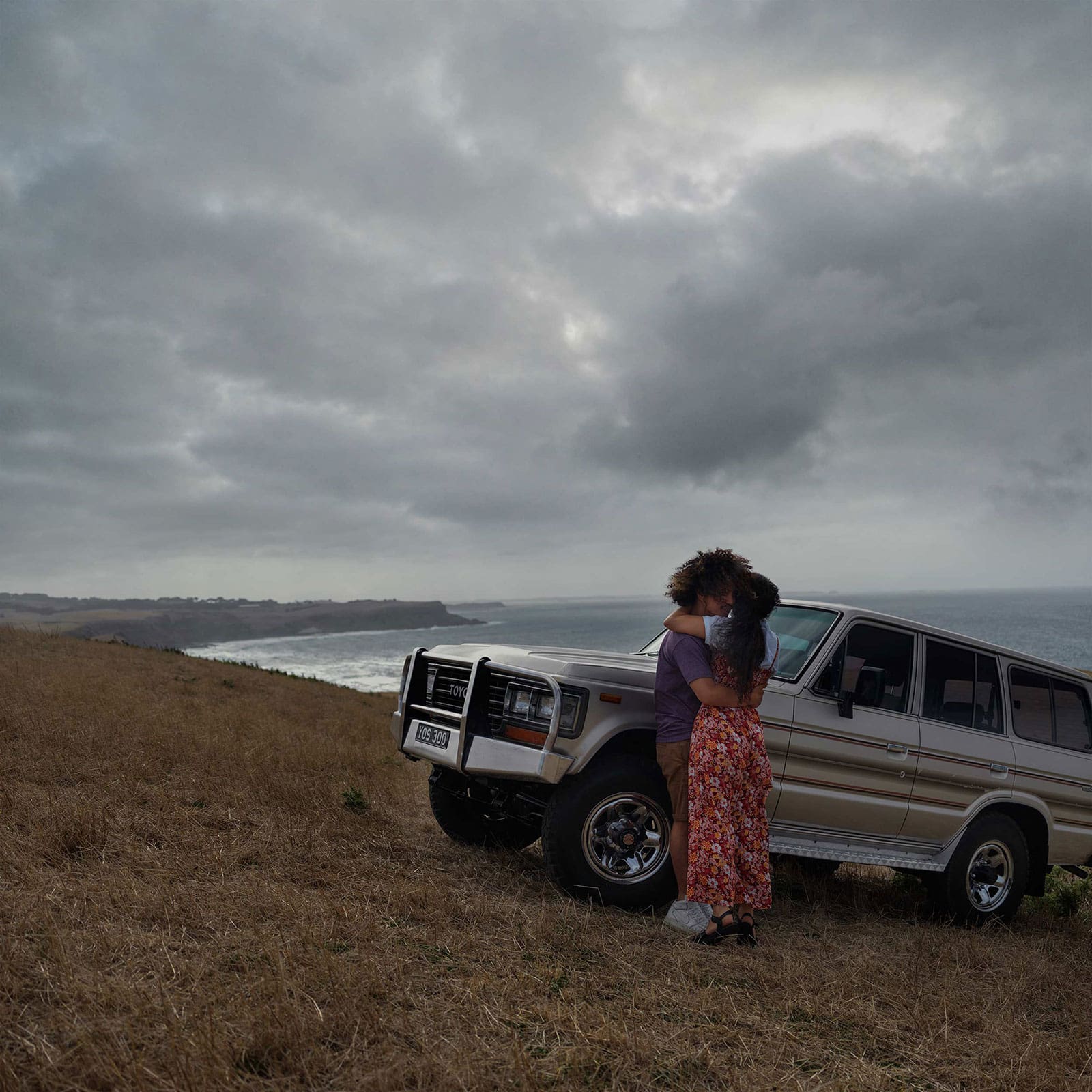 Couple embraces beside a tan SUV on a grassy hill overlooking the ocean under a cloudy sky. Automotive, travel, and lifestyle imagery.