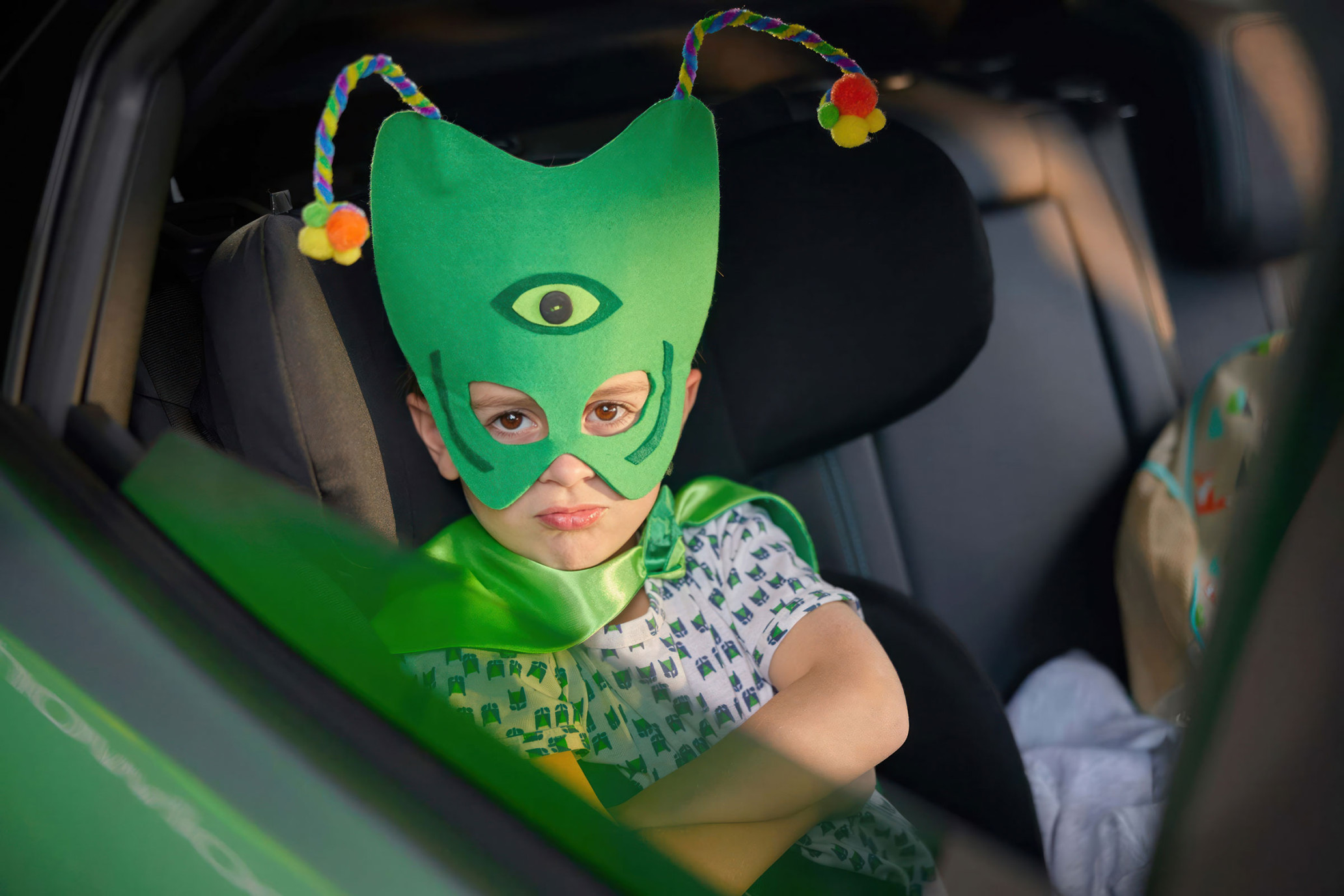 Young child in green costume mask with cape, posing in car seat with arms crossed, looking at the camera with a serious expression.