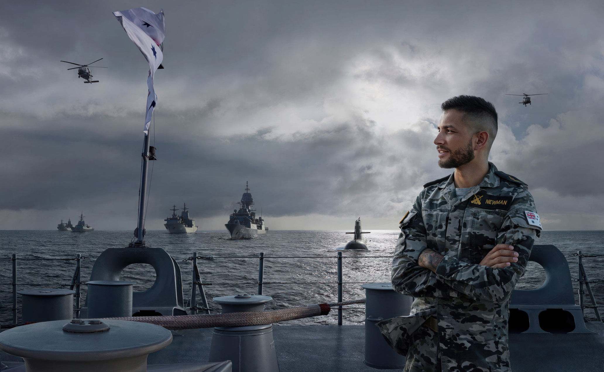 Man in military uniform on deck of naval ship with warships, helicopter, and submarine on the water under a cloudy sky.