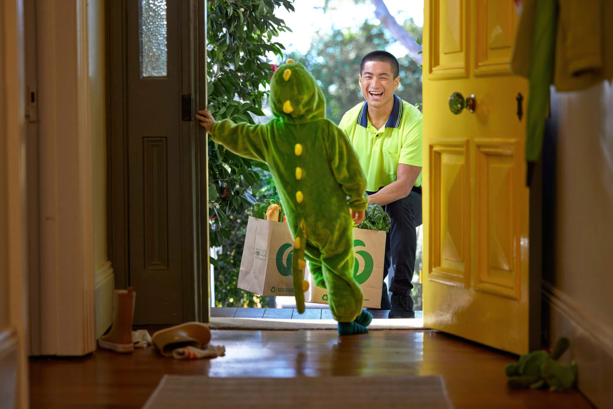 Child in a dinosaur costume receives groceries from a delivery person at the front door of a home with a yellow door.