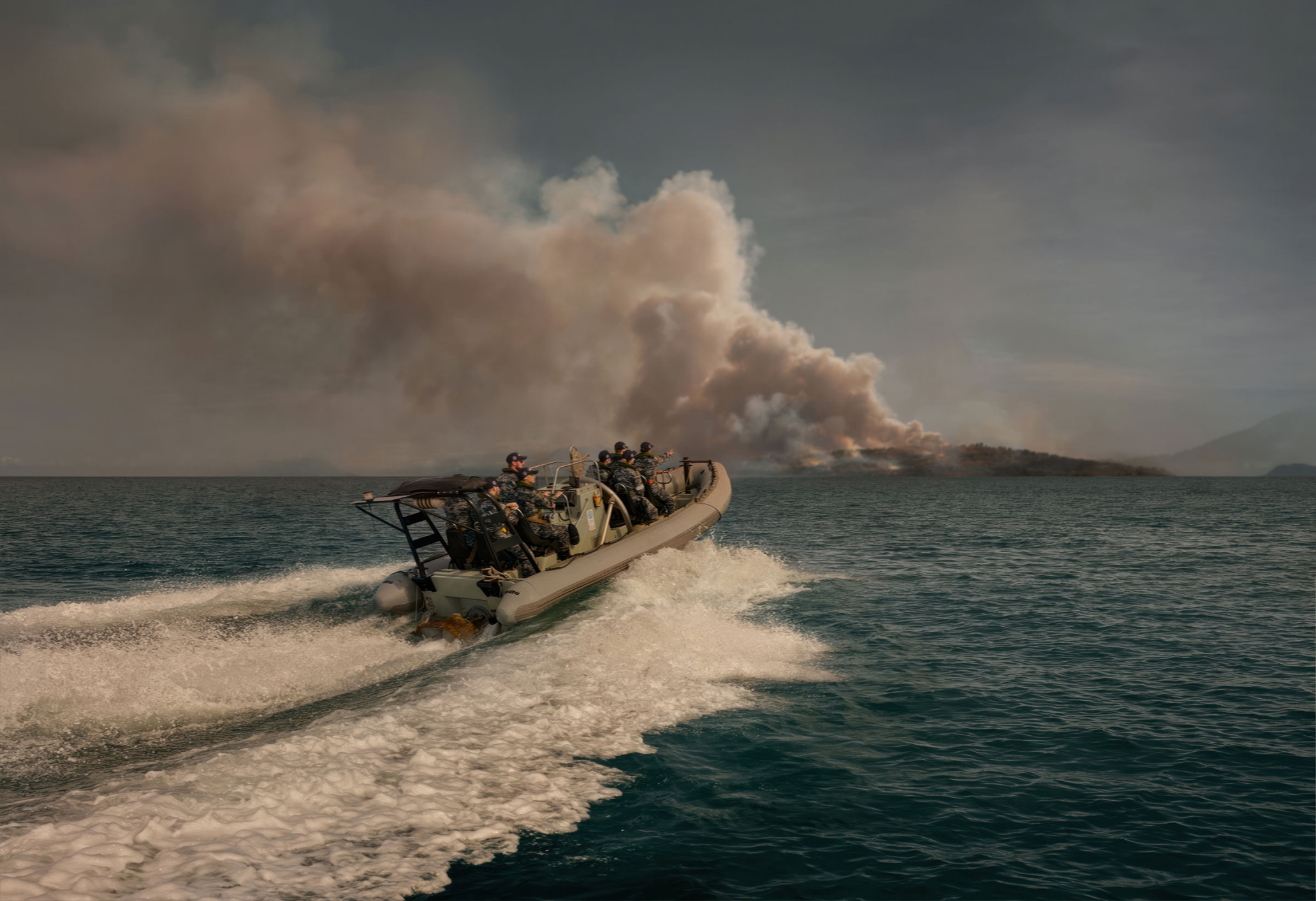 Speedboat carrying group of people moving fast on water with island with fire and smoke in background on overcast day.