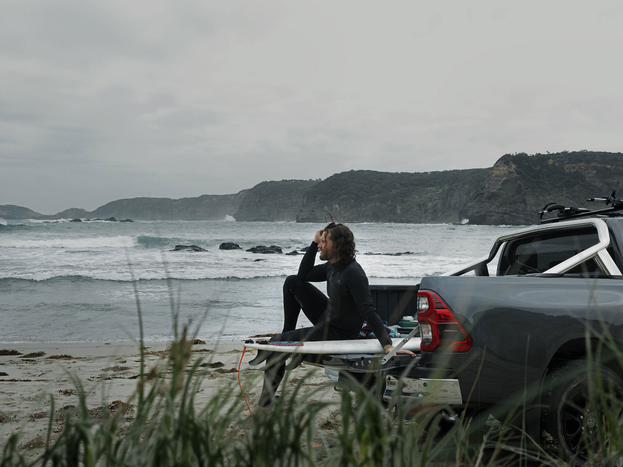 Male surfer sits on surfboard in the bed of a pickup truck near a beach on a cloudy day with an ocean and headland background.