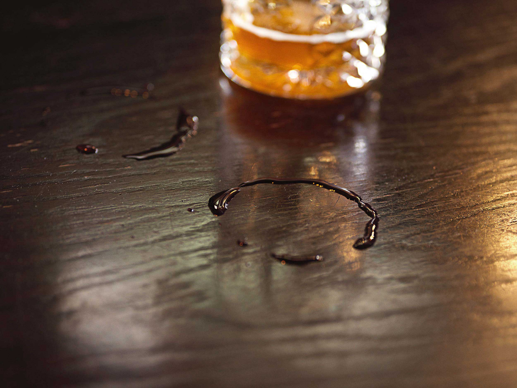Close-up on the surface of a dark wooden table with a ring-shaped liquid stain and a partially visible glass filled with a light-colored liquid.