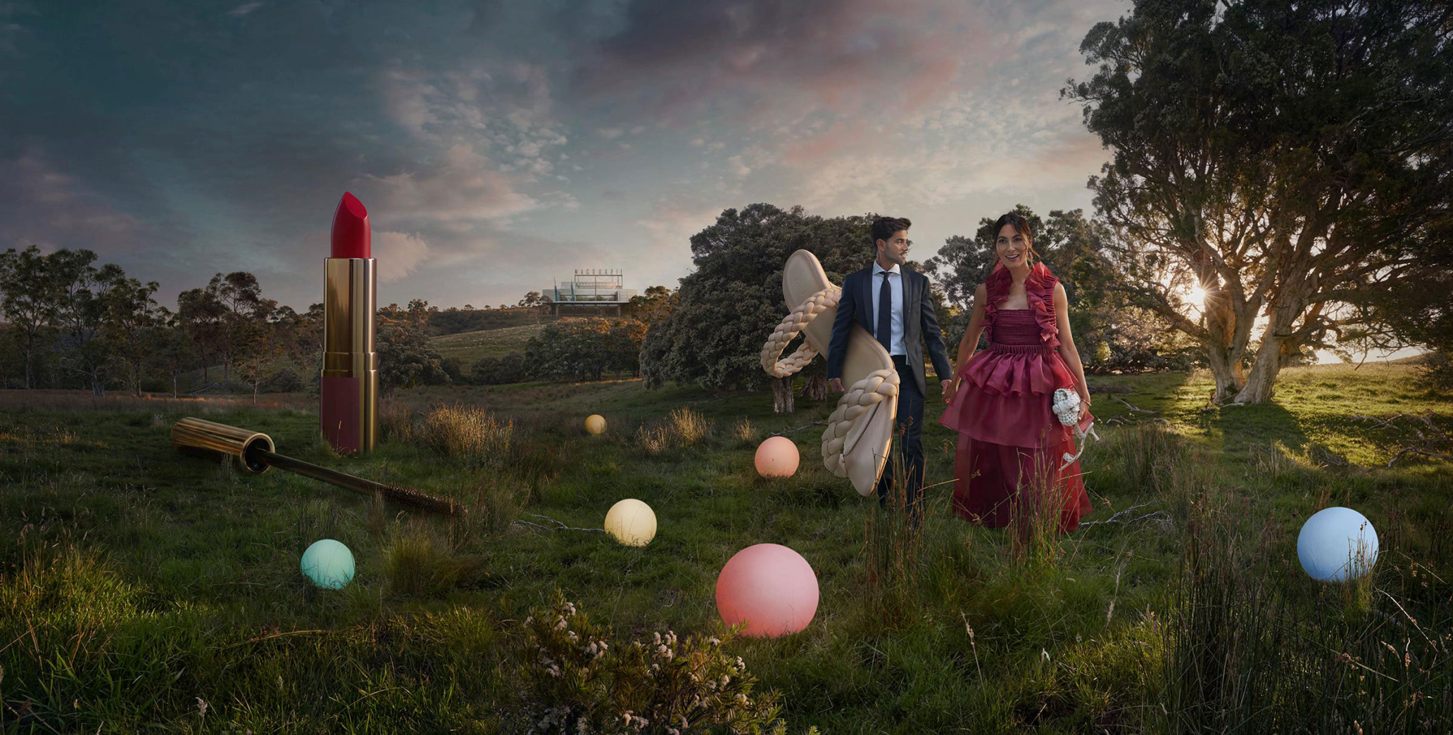 Couple in formal wear walking in grassy field alongside oversized lipstick, mascara, and decorative spheres under dramatic sky.