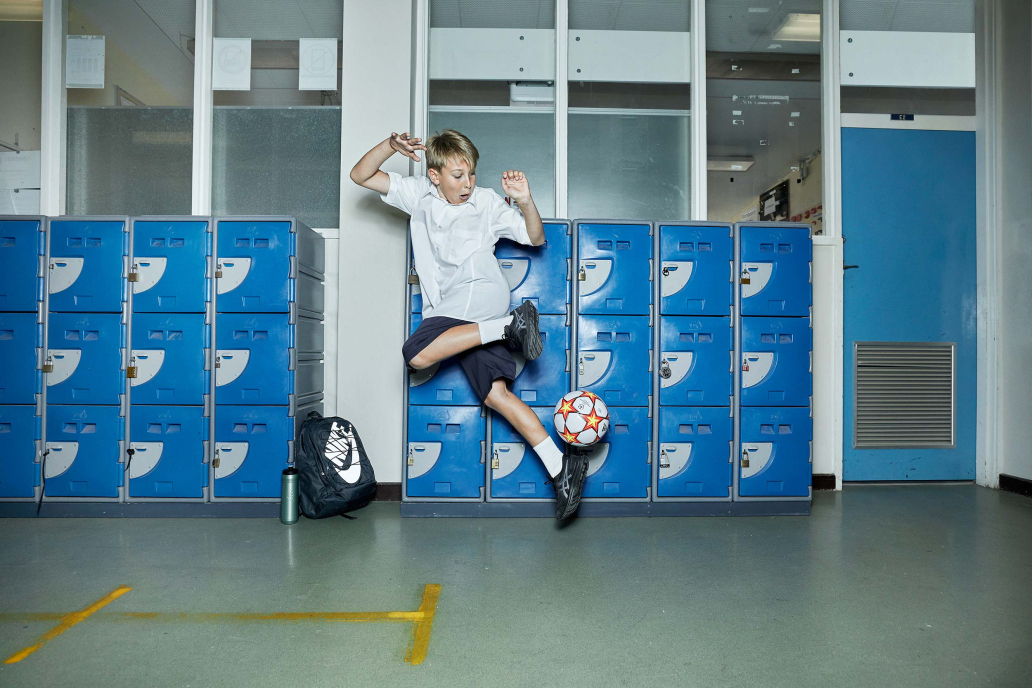 Young boy in mid-air kicks soccer ball in school hallway with lockers, backpack, uniform, and casual shoes, action shot, indoor.
