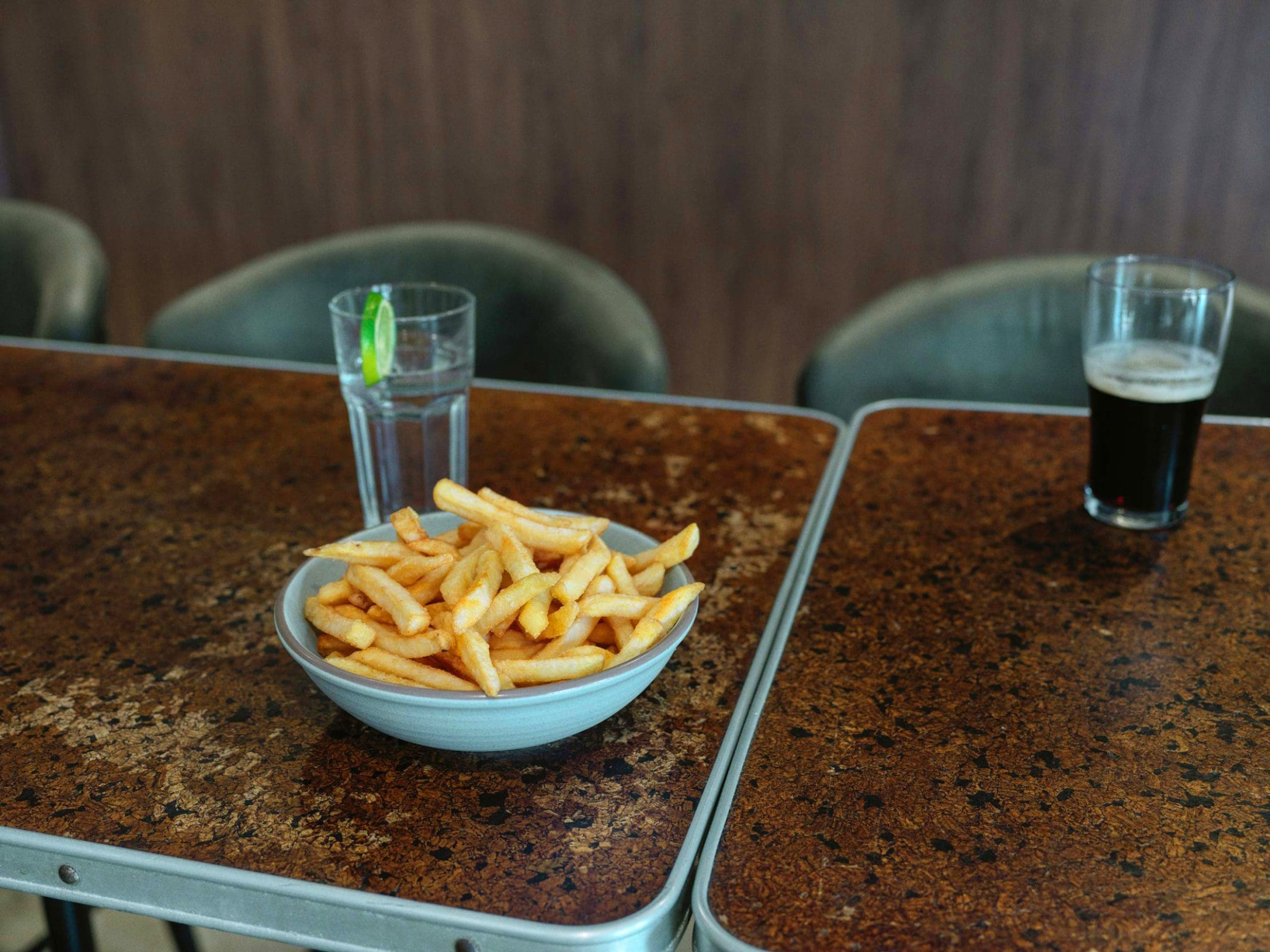 Bowl of french fries on a dark brown speckled table next to a glass of water with lime and dark beverage in a glass.