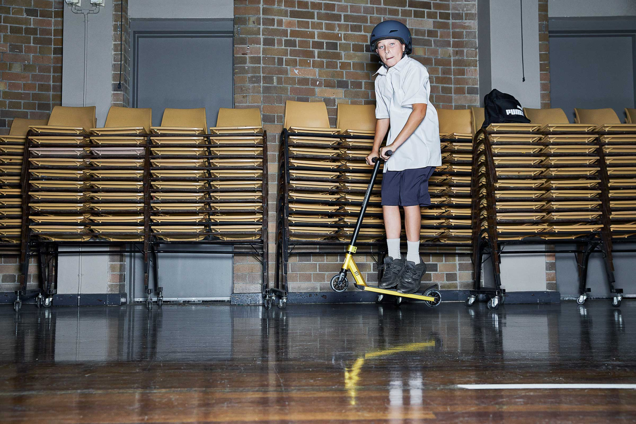 A young person in a school uniform and helmet rides a scooter indoors, with rows of stacked chairs in the background.