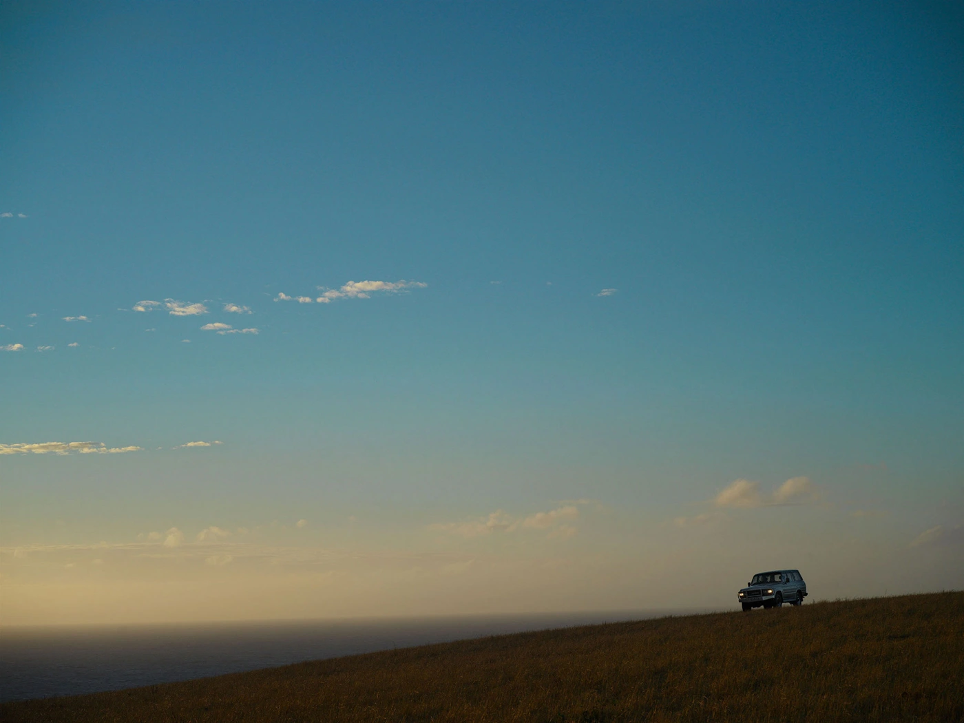 A vintage SUV driving along a grassy ridge under a vast clear blue sky during sunset, photographed by Andreas Smetana.