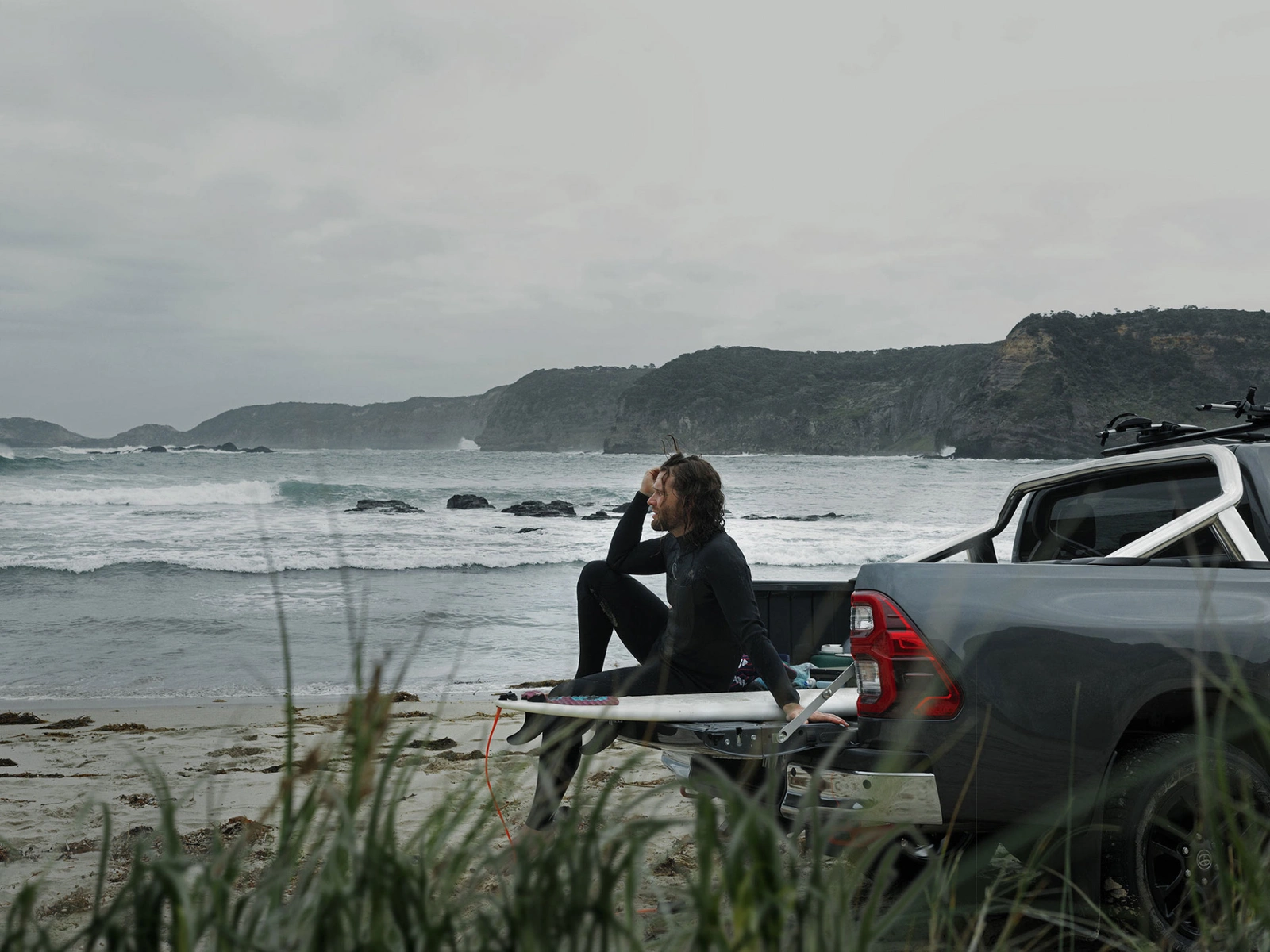 A man in a wetsuit sits on a surfboard in the back of a pickup truck parked on a sandy beach overlooking the ocean.