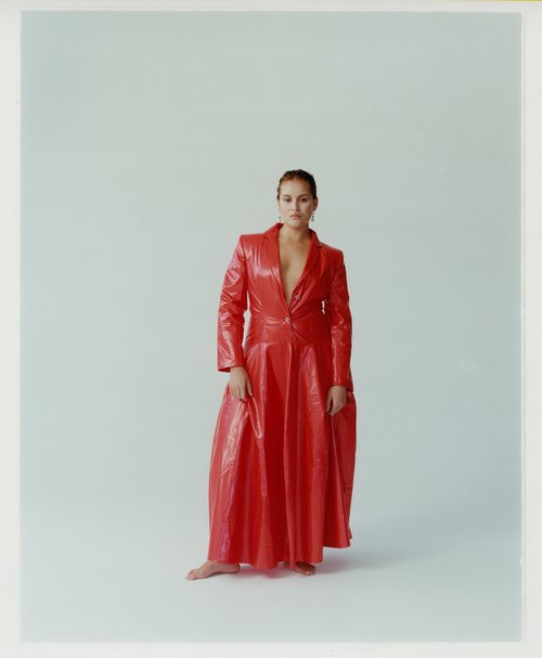 A woman stands in a shiny red blazer-dress, bare feet, against a light background in a studio. The dress has lapels and buttons.