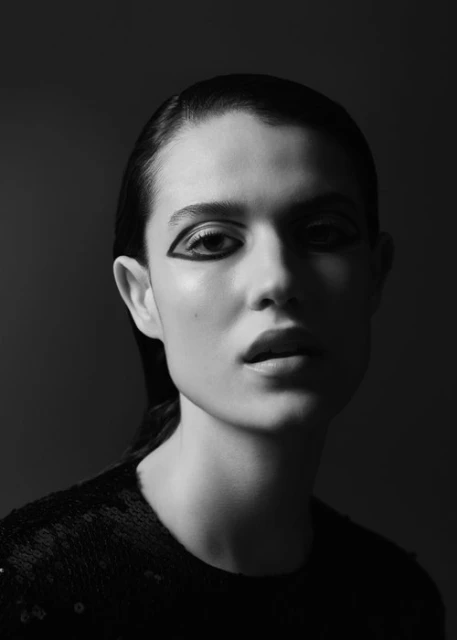 Black and white portrait of a young woman with dark hair and bold black eyeliner, against a dark gray backdrop, in a beauty shot.