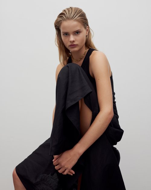 Fashion model in a black dress poses in a studio with soft lighting. The model has blonde hair and is wearing gold necklace.