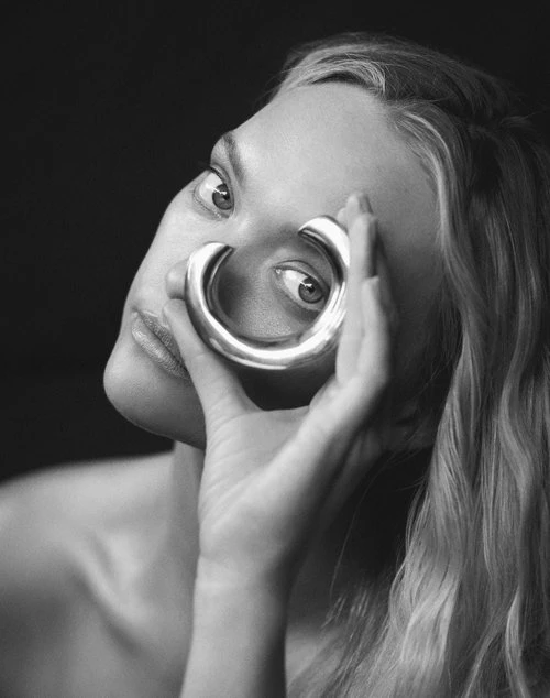 Black and white portrait of a fair-skinned woman holding a silver cuff bracelet up to her eye in front of a dark, stark background.