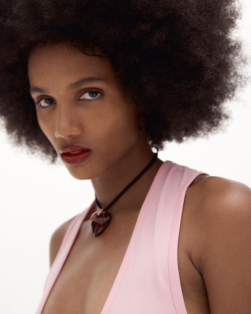 Close-up portrait of an African-descent adult woman with dark hair, wearing a pink top and heart-shaped necklace against a white background.