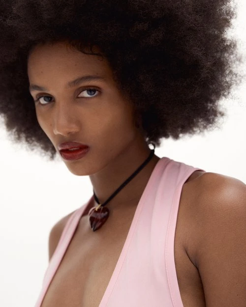 Close-up portrait of an African-descent adult woman with dark hair, wearing a pink top and heart-shaped necklace against a white background.