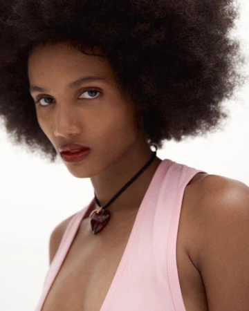 Close-up portrait of an African-descent adult woman with dark hair, wearing a pink top and heart-shaped necklace against a white background.
