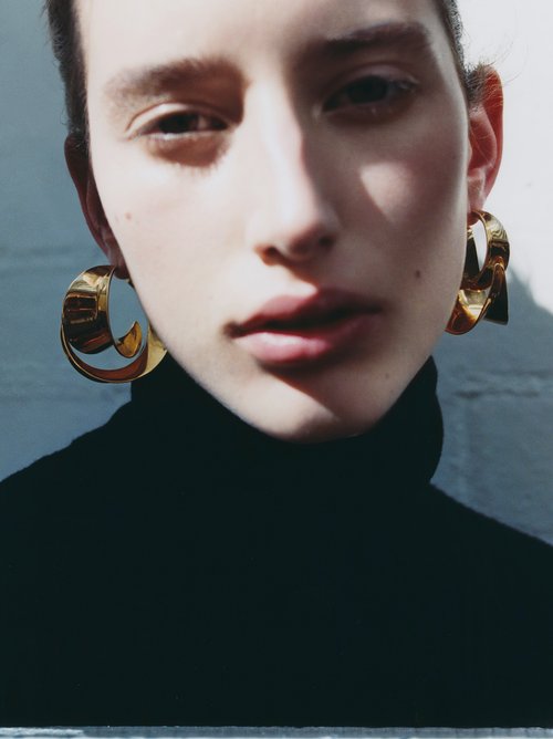 Close-up portrait of a young adult female model wearing gold earrings and a black turtleneck sweater against a textured white background.