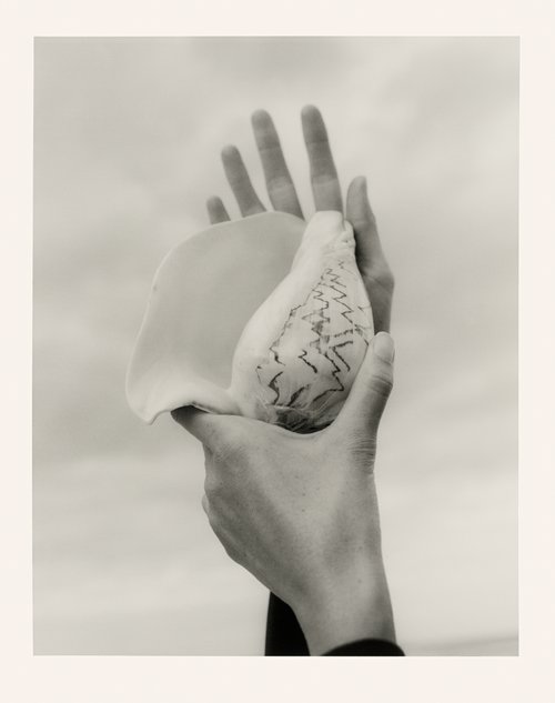 Surreal black-and-white close-up photograph of two hands holding a large conch shell against a cloudy sky, artful and conceptual.