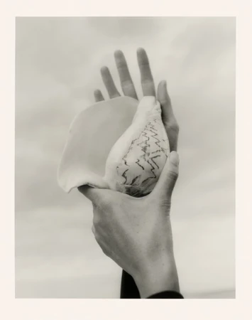 Surreal black-and-white close-up photograph of two hands holding a large conch shell against a cloudy sky, artful and conceptual.