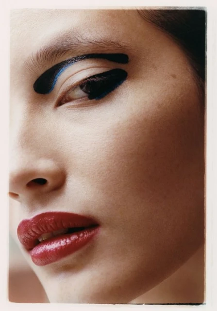 Close-up portrait of a woman with bold black and blue makeup on her eyebrow and eyelid, paired with glossy red lipstick.