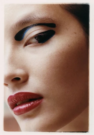 Close-up portrait of a woman with bold black and blue makeup on her eyebrow and eyelid, paired with glossy red lipstick.