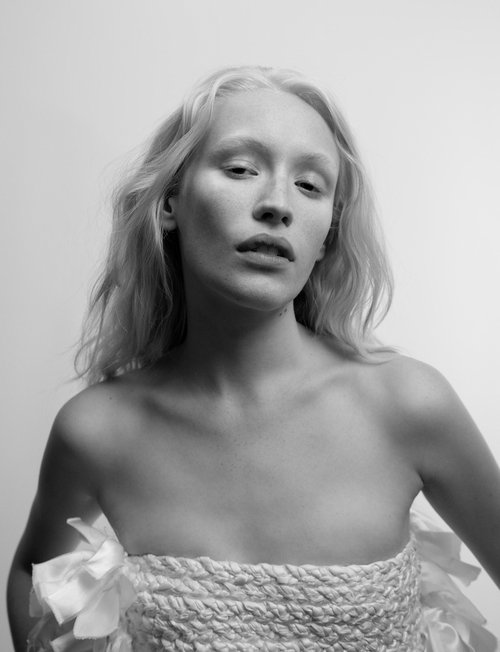 Monochrome portrait of a young woman with fair skin and blonde hair, wearing a woven top with frills against a white backdrop.