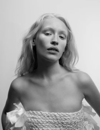 Monochrome portrait of a young woman with fair skin and blonde hair, wearing a woven top with frills against a white backdrop.
