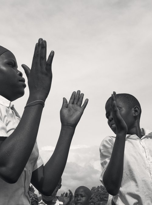 Black and white photograph of African children outside with their hands raised against a cloudy sky, appearing to be clapping or singing.