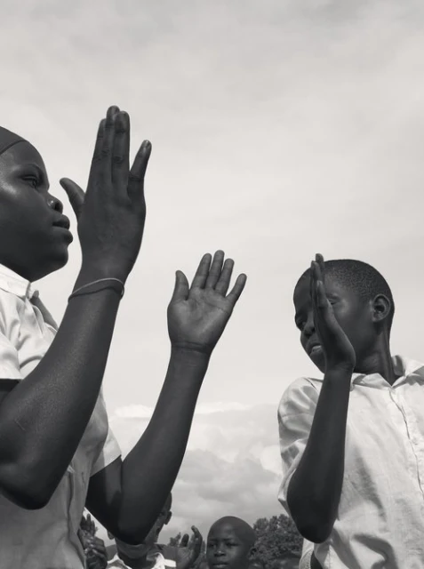 Black and white photograph of African children outside with their hands raised against a cloudy sky, appearing to be clapping or singing.