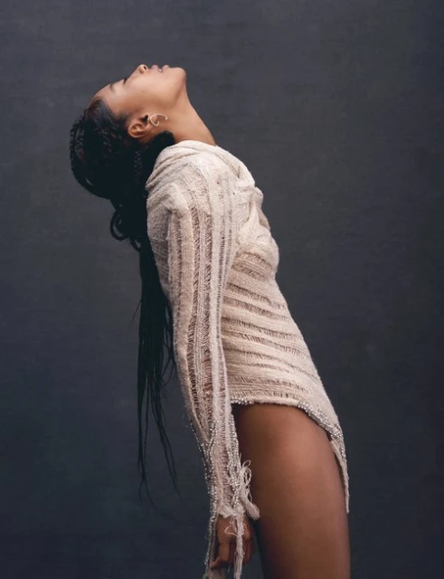 Portrait of a woman with dark hair wearing a textured off-white dress, looking upward in a photo studio with a dark background.